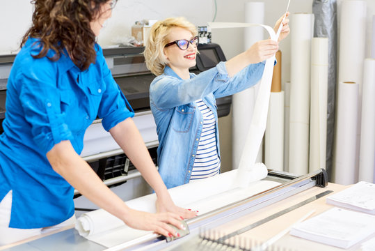 Portrait of two female operators in modern printing shop or publishing company, focus on smiling blonde woman cutting  paper and loading plotter machines, copy space