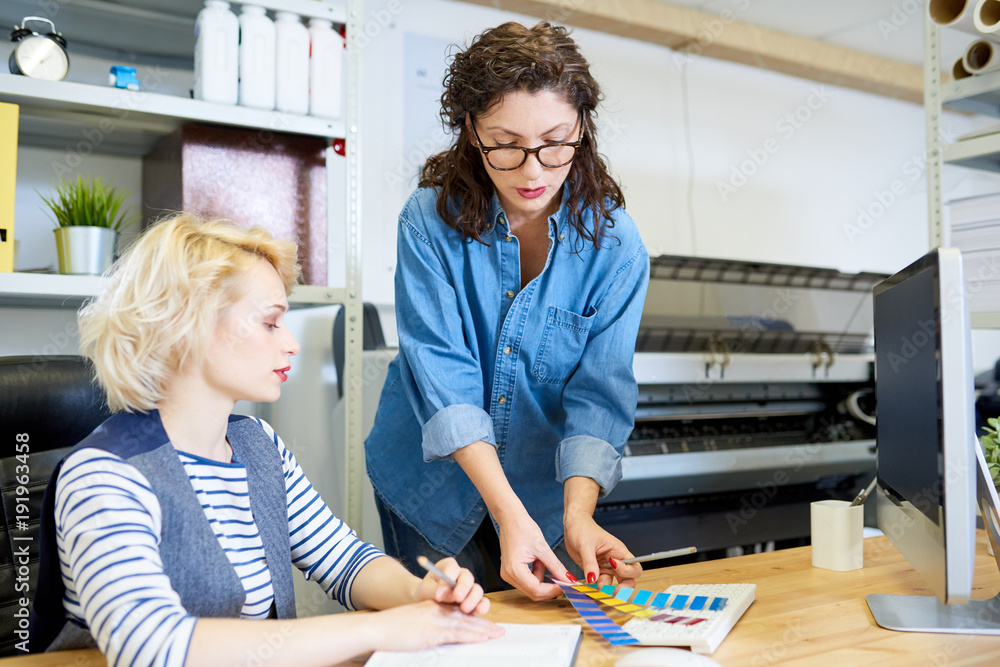 Poster portrait of two graphic designers choosing color palette from swatches while working at desk in prin