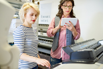 Portrait of two women using plotter machine operating it via digital tablet while working  in...