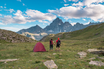 Fototapeta premium Couple looking at the majestic view of glowing mountain peaks at sunset high up on the Alps. Rear view with camping tent, focus in the background, toned and filtered image.