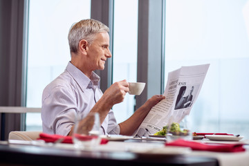 New day. Cheerful optimistic mature man sipping coffee while carrying newspaper and finishing breakfast