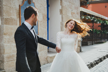 Wedding couple runs in the old city. Blue vintage doors and cafe in ancient sunny town on background. Rustic bride with hair down and groom in grey suit and bow tie. Fun, laugh and happy emotions.