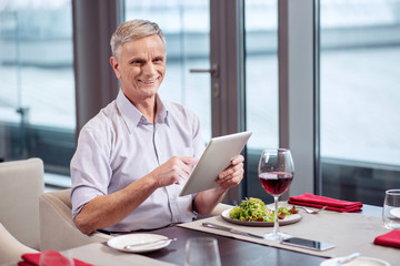 Fototapeta na wymiar Advanced technology. Handsome jolly mature man sitting while holding tablet and grinning at the restaurant