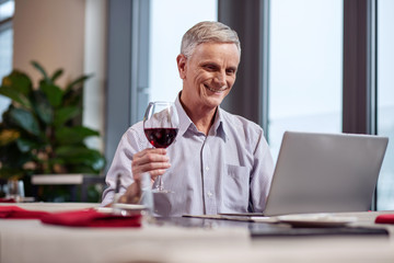 Online communication. Merry handsome mature man drinking wine while smiling and using laptop