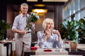 Where you. Musing good looking mature woman holding cup while waiting at the restaurant and staring aside