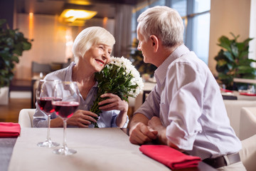 Mature love. Lovely sincere mature woman grinning while holding flowers while man staring at her