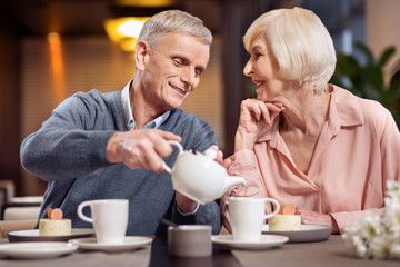 Hot tea. Pleased gay mature man serving tea for woman while smiling and woman staring at him