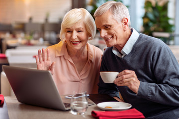 Hi granddaughter. Attractive jolly mature couple sitting at the table and looking at screen while man drinking coffee