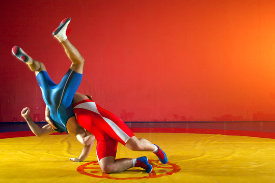 Two young men in blue and red wrestling tights are wrestlng and making a hip throw on a yellow wrestling carpet in the gym