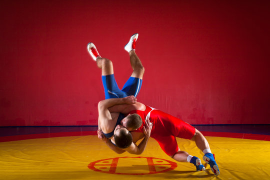 Two young men in blue and red wrestling tights are wrestlng and making a hip throw on a yellow wrestling carpet in the gym
