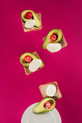 close up view of toasts, avocado pieces, mozzarella cheese and cherry tomatoes falling on plate isolated on pink