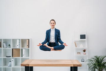 smiling young businesswoman with closed eyes meditating while levitating at workplace - obrazy, fototapety, plakaty