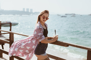 attractive young woman with plastic cup of cocktail on wooden pier in front of sea