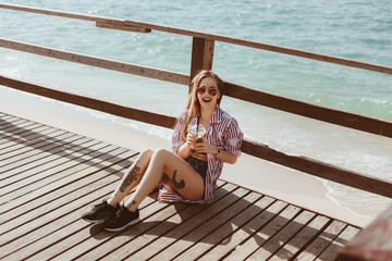 laughing young woman with plastic cup sitting on wooden pier in front of sea