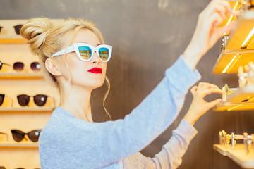 Happy blonde fashionable woman in sunglasses with red lipstick choosing eyeglasses in display of...