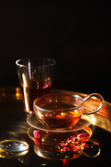 Vertical close-up shot of small glass teapot and cup with hot red tea, dried rose petals, pocket magnifier on golden chain on golden tray. Evening light. Golden color concept.