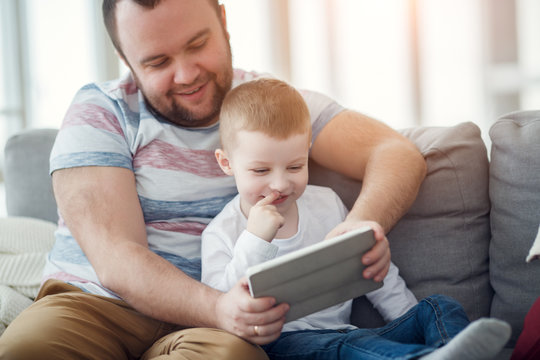 Photo of dad with his son with tablet sitting on gray sofa