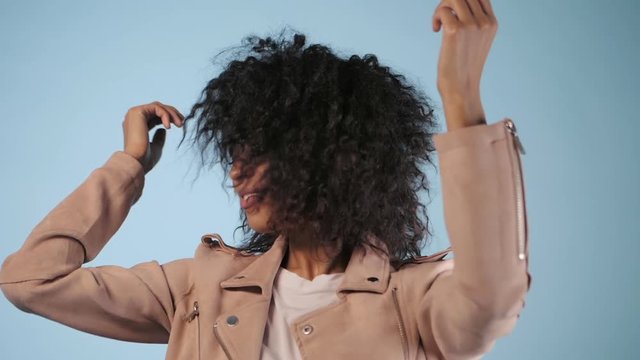 beautiful mixed race woman in pink fashion jacket with afro hair having fun smiling and dancing in studio over blue background. Trendy hipster african girl. Slow motion.