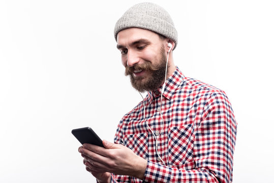 Underground culture. Pleasant bearded man in a grey beanie and a checked shirt standing half-turned and listening to music in headphones while posing isolated on a white background