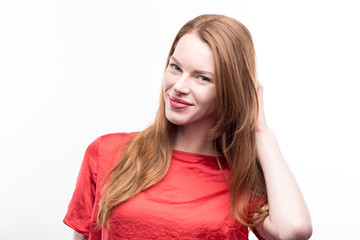 Appealing pose. The portrait of a gorgeous ginger-haired young woman in a red t-shirt posing with her hand in hair while standing isolated on a white background