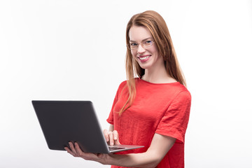 Fototapeta premium Working thoroughly. Beautiful ginger-haired young woman in eyeglasses holding a laptop and grinning at camera while posing isolated on a white background