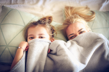 Portrait mother and daughter hide under blanket on a bed.