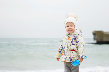 Cute little girl playing on the sandy beach. Happy child wearing warm floral print jacket, pom pom hat and scarf playing outdoors on fall, winter or spring day.