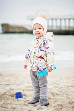 Little girl playing on the sandy beach. Happy child wearing warm floral print jacket, pom pom hat and scarf playing outdoors on fall, winter or spring day.