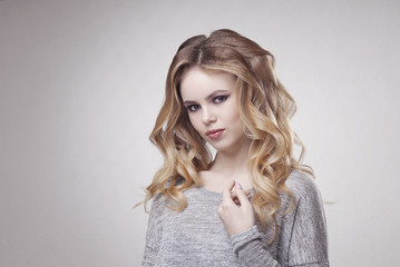 Blonde girl in a gray sweater with long curled hair and makeup Smokey eyes on gray background