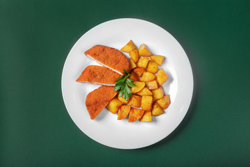 Schnitzel with fried potatoes on a white plate on a green background. View from above. Garnished with greenery.