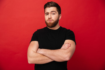 Portrait of muscular serious man 30s wearing black t-shirt posing on camera with hands crossed, isolated over red background