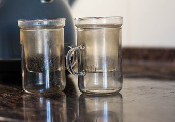 Making tea (green tea leaves) in a glass cup, wit two pieces, green tea leaves with hot water in a transparent clear cup with a kettle near it, making tea with kettle