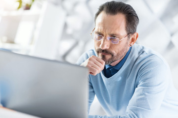 Thoughts. Handsome thoughtful dark-haired bearded man touching his chin and working on his laptop while sitting at the table