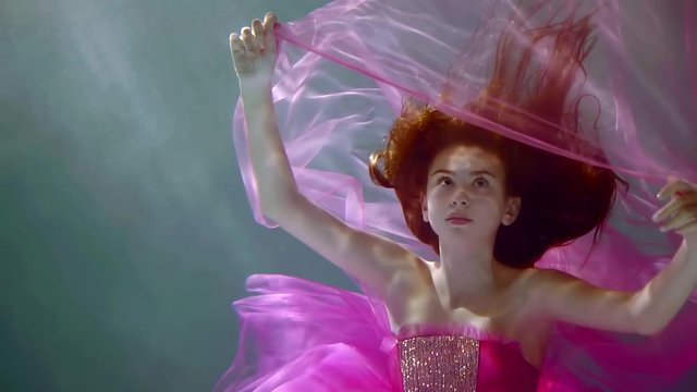Graceful slender young girl model posing underwater in the sun dressed in fuchsia color dress