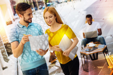 Important document. Clever friendly experienced colleagues standing outdoors and looking at the important document while being in a lovely cafe with their friend sitting behind them