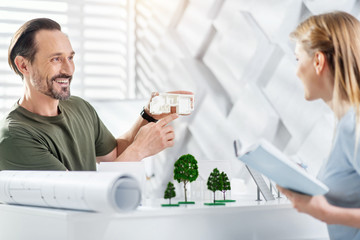 Discussing project. Attractive exuberant bearded man smiling and holding a house miniature while working on the project with his co-worker