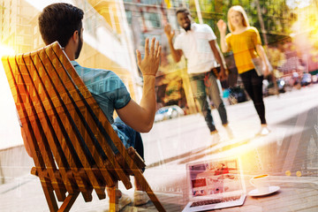 Waving hands. Calm young man having a good time in a comfortable armchair outdoors and waving to his friends when noticing them coming to him
