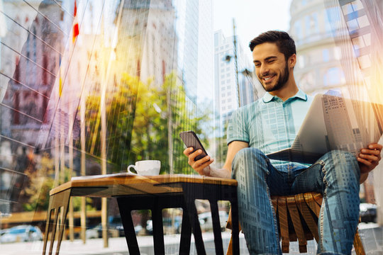 Feeling happy. Cheerful optimistic young man having a good time in a lovely cafe outdoors with a modern laptop on his knees and smiling while looking at the screen of his amazing smartphone