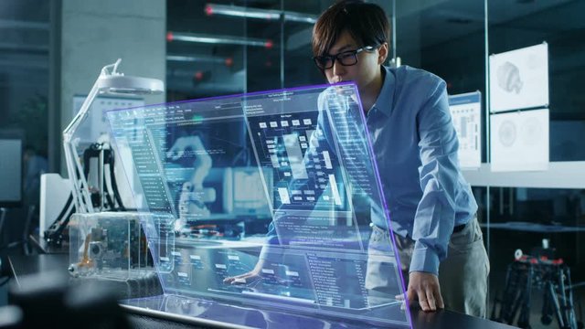 Asian Neural Microchip Design Engineer Uses Modern Computer With Transparent Holographic Display. Monitor Shows Advanced Technology Interface . Shot in Modern Glass and Concrete Office.