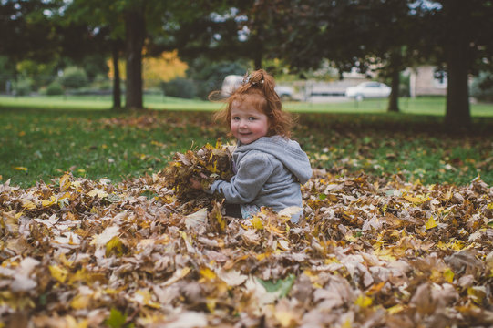 Portrait of red haired female toddler in park with bundles of autumn leaves