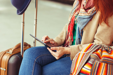 Hipster tourist texting message on tablet or technology mockup. Person traveler using computer on train station background close. Female hands holding gadget on blurred backdrop. Luggage