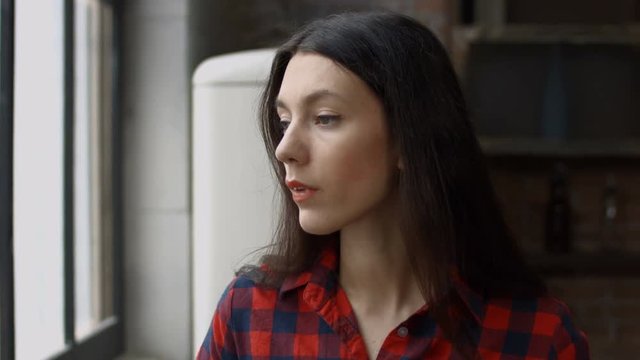 Portrait of pensive attractive woman in causal outfit drinking fresh blended detox smoothie while looking out window in domestic kitchen. Thoughtful young housewife enjoying healthy drink at home.