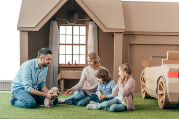 beautiful young family sitting on yard of cardboard house with their puppy
