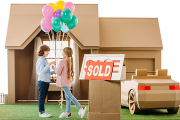 kid presenting balloons to girlfriend in front of cardboard house with sold signboard on foreground...