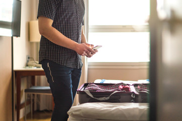Man using smartphone in hotel room with open baggage and suitcase on bed. Mobile phone and packing....