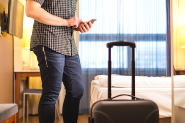 Fototapeta na wymiar Man using smartphone in hotel room with baggage and suitcase. Tourist with mobile phone in holiday rental apartment. Calling taxi cab or doing online check-in. Travel and smart technology concept.