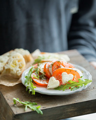 Hearts of sliced Italian cheese Mozzarella with a fresh arugula, tomatoes, olive oil, pesto sauce and bread toasts on a wooden box.   Atmospheric romantic food photo with hands  Restaurant concept.