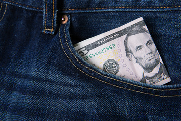 Closeup of the front pocket of faded denim blue jeans with five dollar bills sticking out.