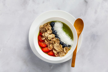 a bowl of smoothies, with cereals for a healthy breakfast and berries, chia seeds and coconut chips freshly sliced fruit, Wheat bran and Cornflake. Served with a wooden spoon on a white marble table