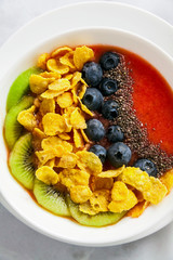 a bowl of smoothies, with cereals for a healthy breakfast and berries, chia seeds and coconut chips freshly sliced fruit, Wheat bran and Cornflake. Served with a wooden spoon on a white marble table
